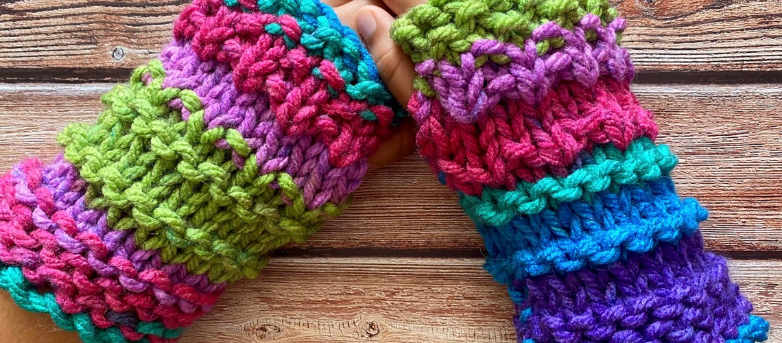 a pair of super chunky wrist warmer fingerless mittens worn on a pair of hands showing the mitts in the knitting pattern
