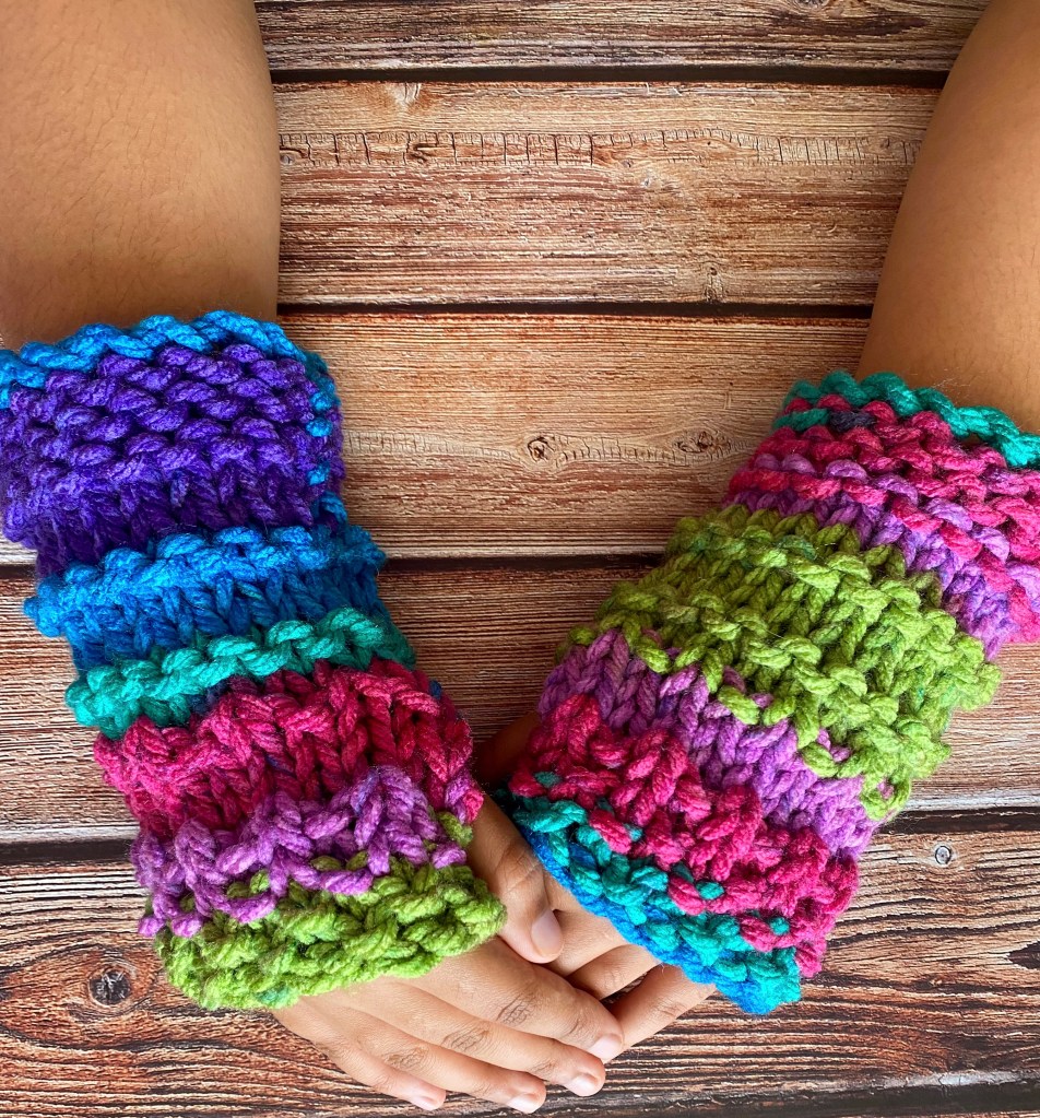 a pair of super chunky wrist warmers in colours of blue, pink and green, showing the mittens from the knitting pattern worn on hands.